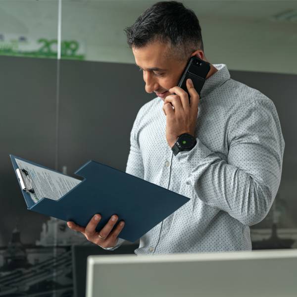 Мъж служител в отдел логистика облечен в сива риза разговаря по мобилен телефон докато разглежда документ в папка